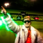 A student wearing sunglasses and a shaggy white fur coat points upward as they DJ at an electronic dance music event. 