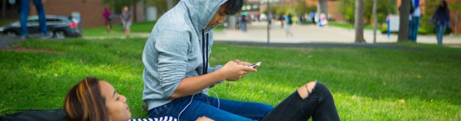 Two students sit in the grass outside on North Campus. One is looking at a smartphone. 