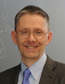 John Ringland smiling. He has short hair and is wearing glasses and a suit and tie. 
