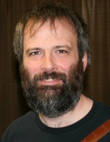Ethan Blanton smiling with a long beard, shirt, and bag strap on his shoulder. 