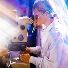 Sideview of a student looking into a microscope with bright streaks of light on the left side of the photo. 