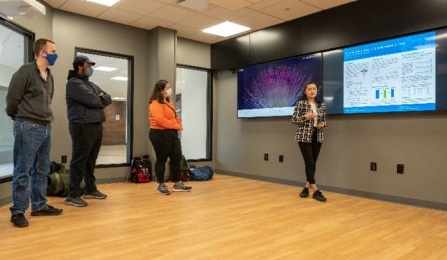 Yihe Yu, a graduate student stands in front of a screen featuring her research and looks out toward the audience while four observers stand to her right slightly in front of, or behind her. 