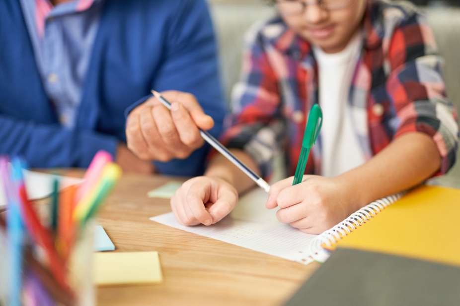 A closeup of a child's hands writing at a desk guided by an adult. 