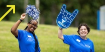 Zoom image: Two UB students wearing UB t-shirts one with a blue foam finger and the other waving a blue and white pom pom 