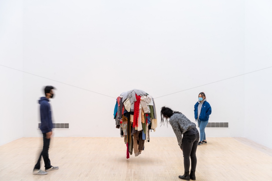three students in an art gallery around a suspended pile of clothing. 