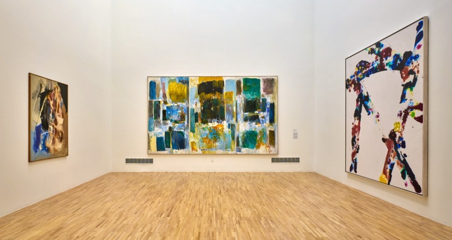 Installation view featuring three works from the UB Art Galleries Collection. From left to right: a color field painting with concentric bands of vibrant hues on a square canvas; a monochromatic abstract composition dominated by vertical black brushstrokes on white ground; and a hard-edged geometric painting with overlapping triangular forms in blue, red, and black. All three works are framed and hung on a white wall in a well-lit gallery space with a concrete floor. The image emphasizes the diversity of modern and contemporary abstraction within the collection. 