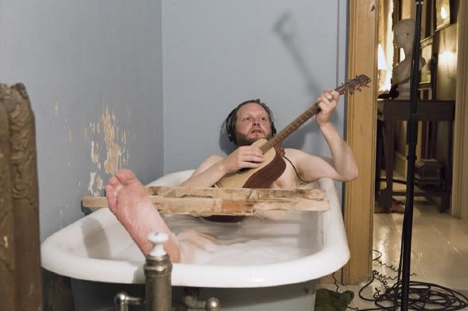 Still Image from Video by Ragnar Kjartansoon of a man with headphones in a bubble bath playing guitar with his right leg stretched out of the bath tub. 