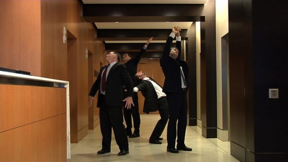 Photograph of four male figures in black suit stretching in the office hallway. 