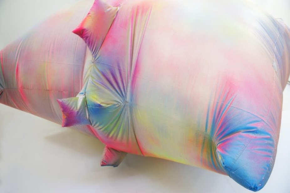 A photo of an inflatable sculpture by Claire Ashley. Only one corner of the piece is in the shot but it is rainbow colored and pillow shaped. 