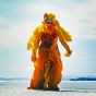 Still image from video by Antonia Wright taken from low angle showing the horizon of the sky blue beach with figure wearing yellow, orange costume standing in the center. 
