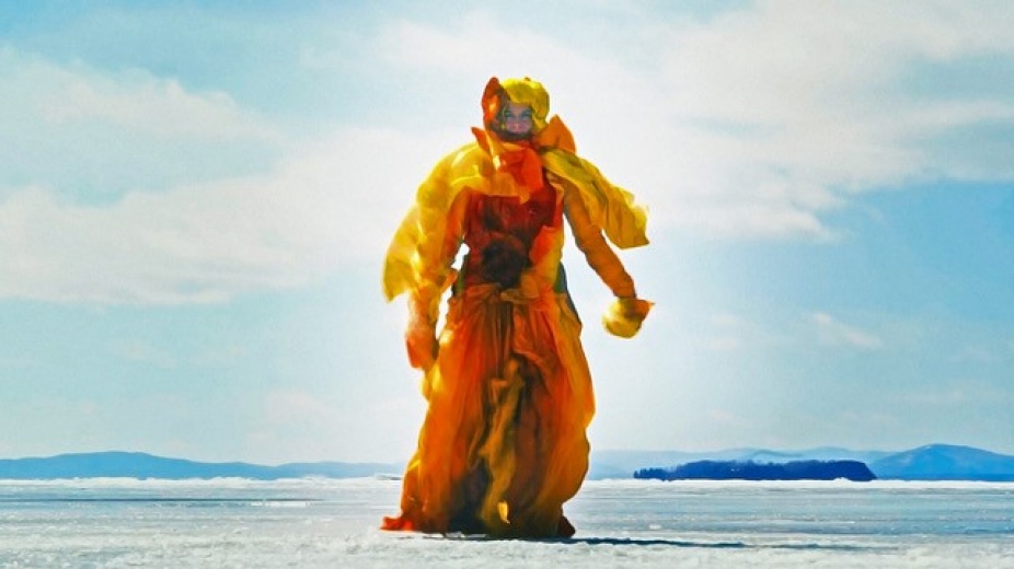 Still image from video by Antonia Wright taken from low angle showing the horizon of the sky blue beach with figure wearing yellow, orange costume standing in the center. 