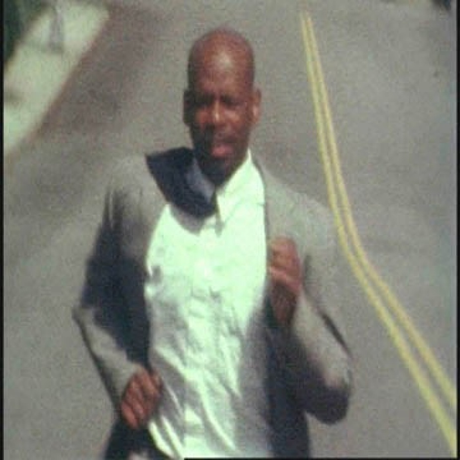 Still Image from a video by Jefferson Pinder from the video "Marathon." A Black man is running on a road. He wear a gray suit and his tie is flying over his shoulder. 