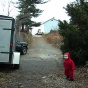 Still image from video by Lenka Clayton. A child with red clothing stands in a driveway next to a trailer and truck and trees. 
