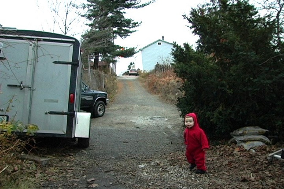Still image from video by Lenka Clayton. A child with red clothing stands in a driveway next to a trailer and truck and trees. 