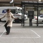 Still Image from Lily McElroy of a woman with a white coat standing in a chalk-drawn square on the sidewalk in the middle of a city. 