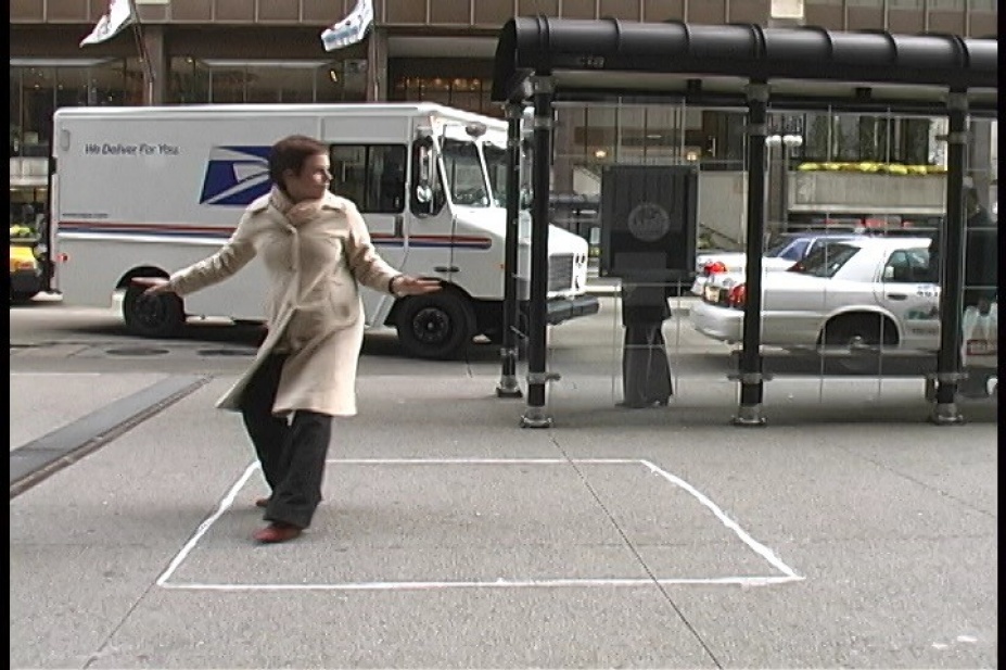 Still Image from Lily McElroy of a woman with a white coat standing in a chalk-drawn square on the sidewalk in the middle of a city. 