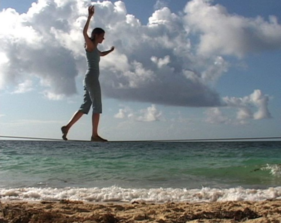 Photography by Janine Antoni of a person with arms rasied walking on a tightrope that is aligning with the horizon of the ocean on the back. 