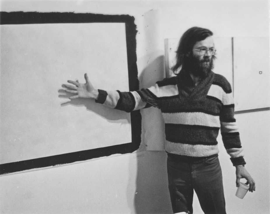 A photograph of Tony Conrad standing in front of a work on a wall. 