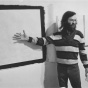 A photograph of Tony Conrad standing in front of a work on a wall. 