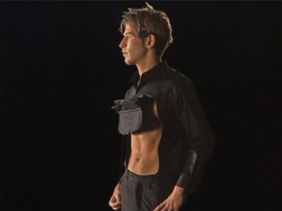 A photo of a man in profile. He wears a black shirt that is pulled up in front, revealing very tight abs. 