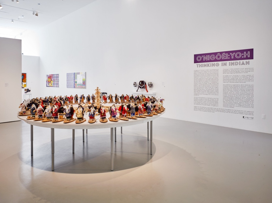 An installation image of Thinking in Indian at UB CFA Gallery. In the foreground is a work by Elizabeth Doxdater where atop large, circular table hundreds of cornhusk dolls are arranged. In the background is a title wall that reads "O’nigöëi:yo:h Thinking in Indian.". 