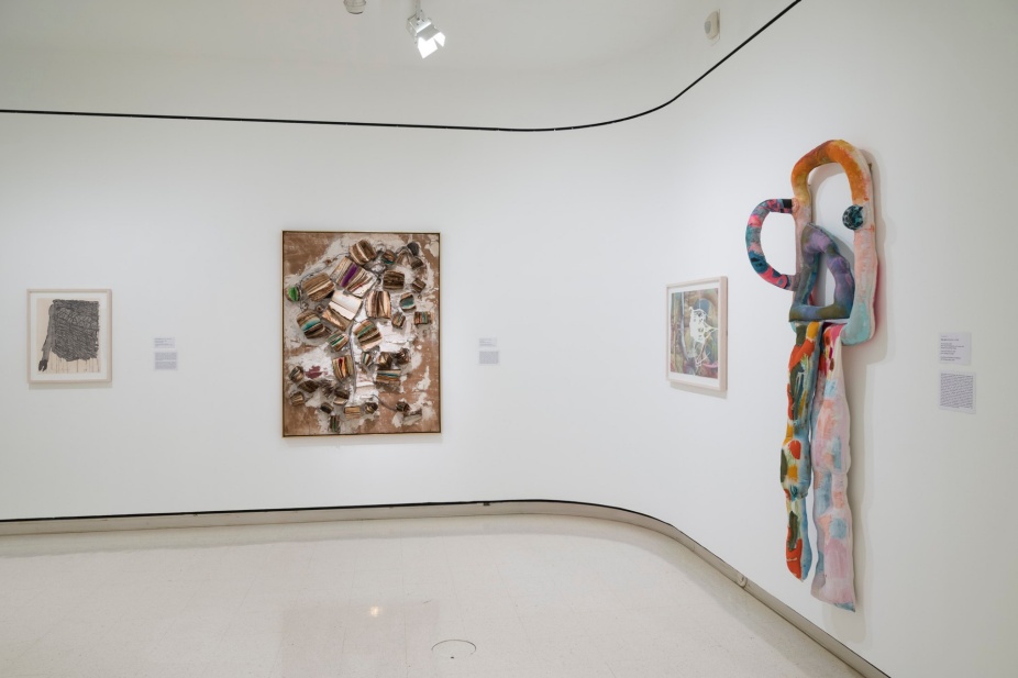 An installation image from In Conversation picturing a corner of a gallery wall. On the left there are two rectanrular works, one mixed media with books affixed to a canvas, and on the right side a recantular work and a big plush fabric sculpture. 