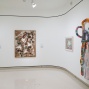 An installation image from In Conversation picturing a corner of a gallery wall. On the left there are two rectanrular works, one mixed media with books affixed to a canvas, and on the right side a recantular work and a big plush fabric sculpture. 