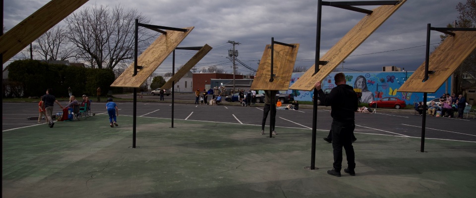 A series of sculptures are installed in a parking lot. The sculptures are about 12feet high, with steel armatures that are mounted in the ground. they have sheets of plywood attached to them at a 45 degree angle that catch the sun. 