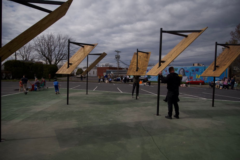 A series of sculptures are installed in a parking lot. The sculptures are about 12feet high, with steel armatures that are mounted in the ground. they have sheets of plywood attached to them at a 45 degree angle that catch the sun. 
