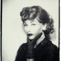Sepia-toned silver gelatin photograph of a woman posed in character. She has glossy, tightly curled hair, dramatic false eyelashes, heavy eyeliner, and dark lipstick. Her chin rests thoughtfully in her gloved hand. She wears a dark-toned garment, possibly a dress or blouse, and gazes forward with a neutral expression&mdash;neither smiling nor frowning. The tightly framed bust portrait highlights the theatrical construction of persona through styling and pose. 