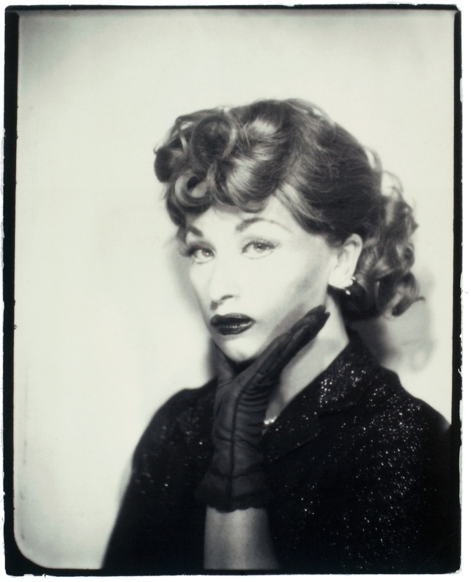 Sepia-toned silver gelatin photograph of a woman posed in character. She has glossy, tightly curled hair, dramatic false eyelashes, heavy eyeliner, and dark lipstick. Her chin rests thoughtfully in her gloved hand. She wears a dark-toned garment, possibly a dress or blouse, and gazes forward with a neutral expression—neither smiling nor frowning. The tightly framed bust portrait highlights the theatrical construction of persona through styling and pose. 