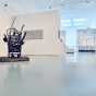 Wide view of an exhibition gallery with mixed-media artworks installed throughout the space. In the foreground, a sculptural assemblage made from found materials sits on a wooden pallet atop a wheeled cart. White partition walls display small grid-based photographs and a gray relief-like wall work. In the background, a large black line drawing installation spells the word &ldquo;CHOOSE&rdquo; across a transparent surface, with additional artworks visible beyond. 