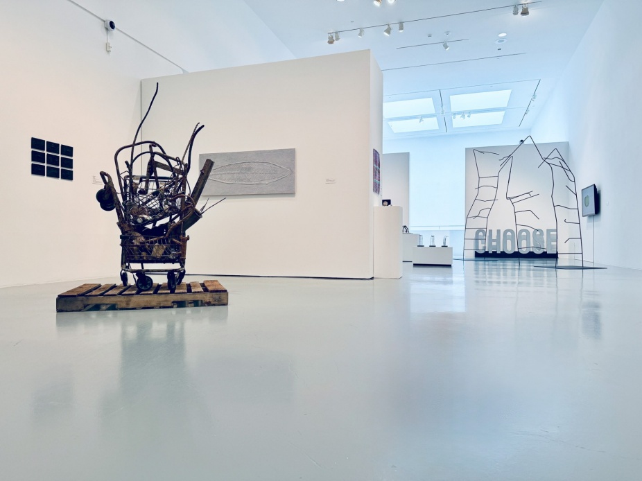 Wide view of an exhibition gallery with mixed-media artworks installed throughout the space. In the foreground, a sculptural assemblage made from found materials sits on a wooden pallet atop a wheeled cart. White partition walls display small grid-based photographs and a gray relief-like wall work. In the background, a large black line drawing installation spells the word “CHOOSE” across a transparent surface, with additional artworks visible beyond. 