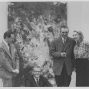 Black-and-white photograph of four people in formal attire posing in front of a large abstract expressionist painting. One man is seated in front wearing glasses and a suit, while the other three&mdash;two men and one woman&mdash;stand behind him. The woman stands with one hand on her hip, smiling at the man beside her, who holds a cigarette. The textured painting behind them features energetic, gestural brushstrokes in light and dark tones. The image evokes a mid-20th century gallery setting. 