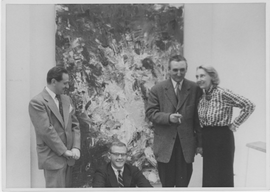 Black-and-white photograph of four people in formal attire posing in front of a large abstract expressionist painting. One man is seated in front wearing glasses and a suit, while the other three—two men and one woman—stand behind him. The woman stands with one hand on her hip, smiling at the man beside her, who holds a cigarette. The textured painting behind them features energetic, gestural brushstrokes in light and dark tones. The image evokes a mid-20th century gallery setting. 