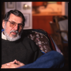 Portrait of an older man with a gray beard and glasses, seated in an armchair indoors. He wears a dark sweater and looks slightly toward the camera, with his hands resting together. The background is softly out of focus, suggesting a lived-in interior space with warm tones. 