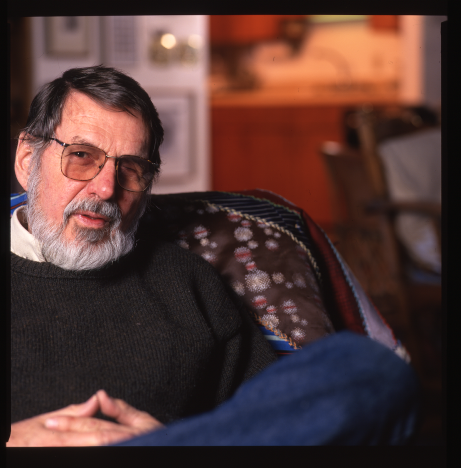 Portrait of an older man with a gray beard and glasses, seated in an armchair indoors. He wears a dark sweater and looks slightly toward the camera, with his hands resting together. The background is softly out of focus, suggesting a lived-in interior space with warm tones. 