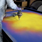 Natvar Bhavsar leans over a large canvas laid flat on a table, sifting dry pigment from a metal funnel onto its surface. The canvas glows with fields of yellow, orange, and violet. The artist wears a pigment-dusted smock and long rubber gloves. 