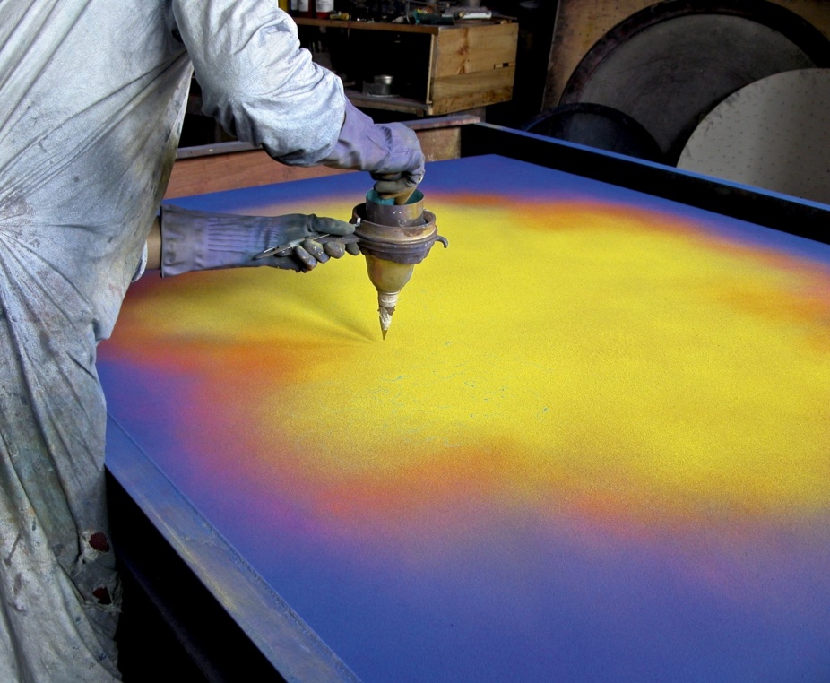Natvar Bhavsar leans over a large canvas laid flat on a table, sifting dry pigment from a metal funnel onto its surface. The canvas glows with fields of yellow, orange, and violet. The artist wears a pigment-dusted smock and long rubber gloves. 