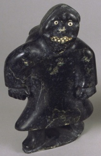 A soapstone sculpture of a figure covered in a garment with two hands and feet poking out. The figure has a toothy smile, white nose, and two wide eyes. 