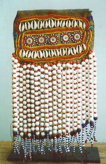 A piece of leather is intricately beaded with small shells, buttons, and beaded fringe. The beads are white, yellow, red, and black. 