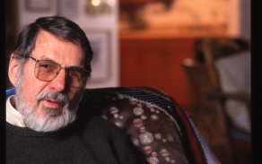 Portrait of an older man with a gray beard and glasses, seated in an armchair indoors. He wears a dark sweater and looks slightly toward the camera, with his hands resting together. The background is softly out of focus, suggesting a lived-in interior space with warm tones. 