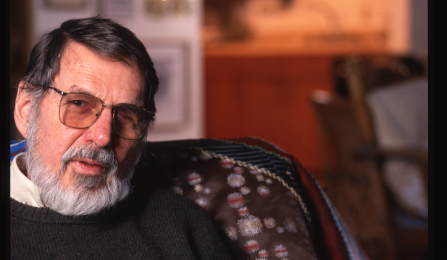 Portrait of an older man with a gray beard and glasses, seated in an armchair indoors. He wears a dark sweater and looks slightly toward the camera, with his hands resting together. The background is softly out of focus, suggesting a lived-in interior space with warm tones. 