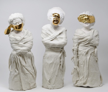 Three porcelain sculptures of women wrapped in draped, straitjacket-like garments stand side by side. Each figure wears a partial gold luster mask that obscures the face. The white porcelain forms are cracked and textured, evoking fragility and constraint. The figures vary in pose, but all appear bound and silent, evoking themes of institutionalization and suppression. 