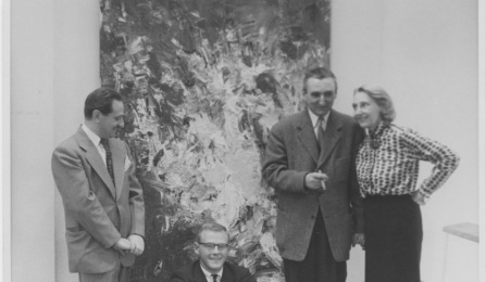 Black-and-white photograph of four people in formal attire posing in front of a large abstract expressionist painting. One man is seated in front wearing glasses and a suit, while the other three&mdash;two men and one woman&mdash;stand behind him. The woman stands with one hand on her hip, smiling at the man beside her, who holds a cigarette. The textured painting behind them features energetic, gestural brushstrokes in light and dark tones. The image evokes a mid-20th century gallery setting. 