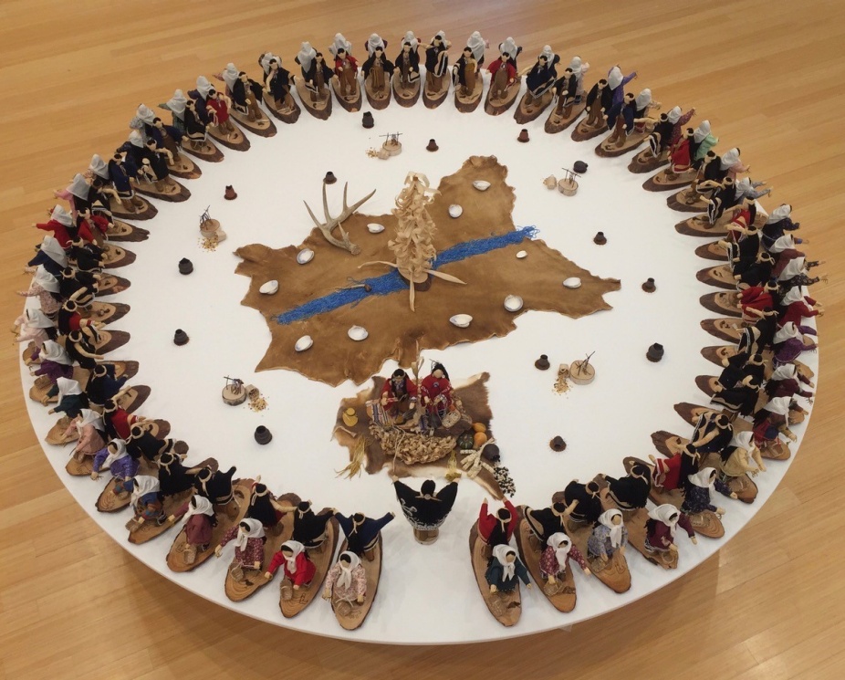 A photo of a work by Elizabeth Doxtater. On a white round table a circle of corn husk dolls in boats faces outward. In the center of the circle is a cow hide and more corhnusk dolls arranged in ceremony. 