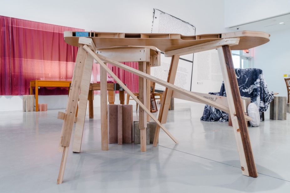 An installation photo of F: Reconstituting Tolstoy College. The central work is a table-like structure configured of many different kinds of legs and supports. 