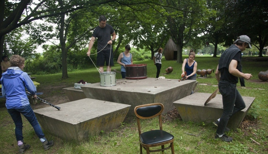 A photograph of people with musical instruments moving around a concrete sculpture. 