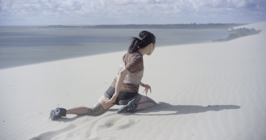 A film still from Eglė Budvytytė, Songs from the Compost: Mutating Bodies, Imploding Stars where a female figure is trying to run while one leg is fully submerged in sand. 