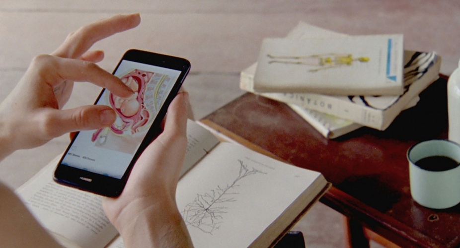 Two fingers pinch the screen of a mobile phone showing an anatomical drawing of a near-term baby, head down in a uterus. The white person’s other hairy hand holds the phone over an open book showing an inverted tree-like chart. Three anatomy and botany books are piled on a small nearby wooden table, which also holds a white cup of dark liquid. 